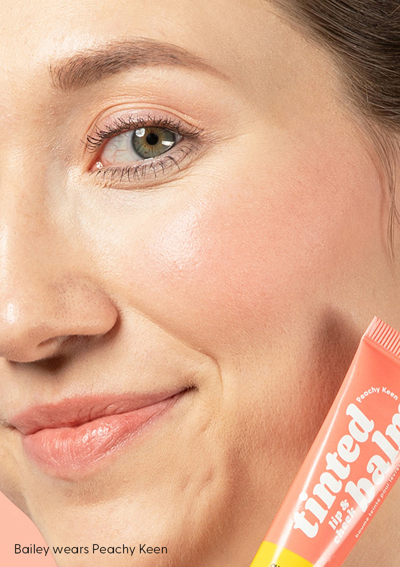 Bailey smiles with AvryBeauty’s Peachy Keen Lip & Cheek Tinted Balm applied to her lips, cheeks, and eyelids, while holding up its vegan skincare tube in front of a peach-colored background.