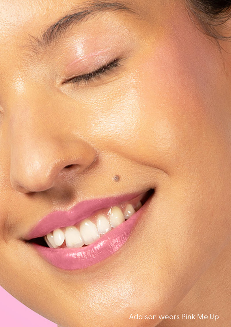 Addison smiles with closed eyes as AvryBeauty’s Lip & Cheek Tinted Balm is applied to her lips, cheek, and eyelid, with a pink backdrop behind her.