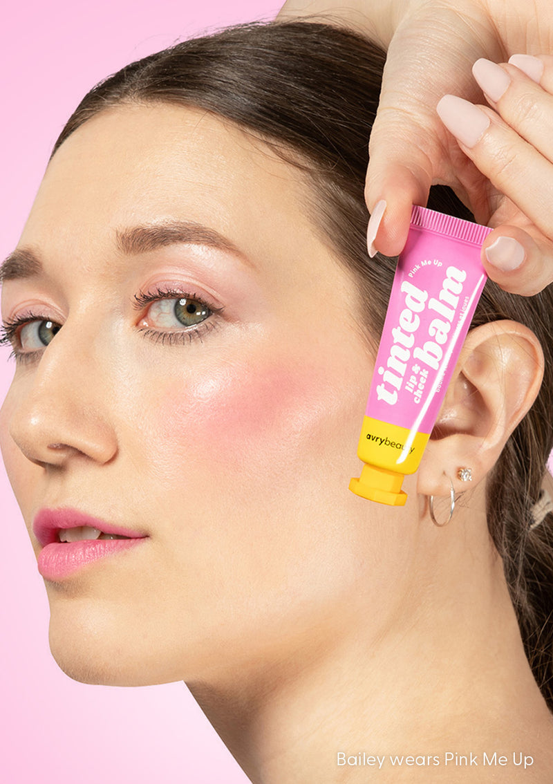 Bailey wears Pink Me Up Lip & Cheek Tinted Balm on her lips, cheeks, and eyelids, holding the tube up to her face against a pink backdrop.