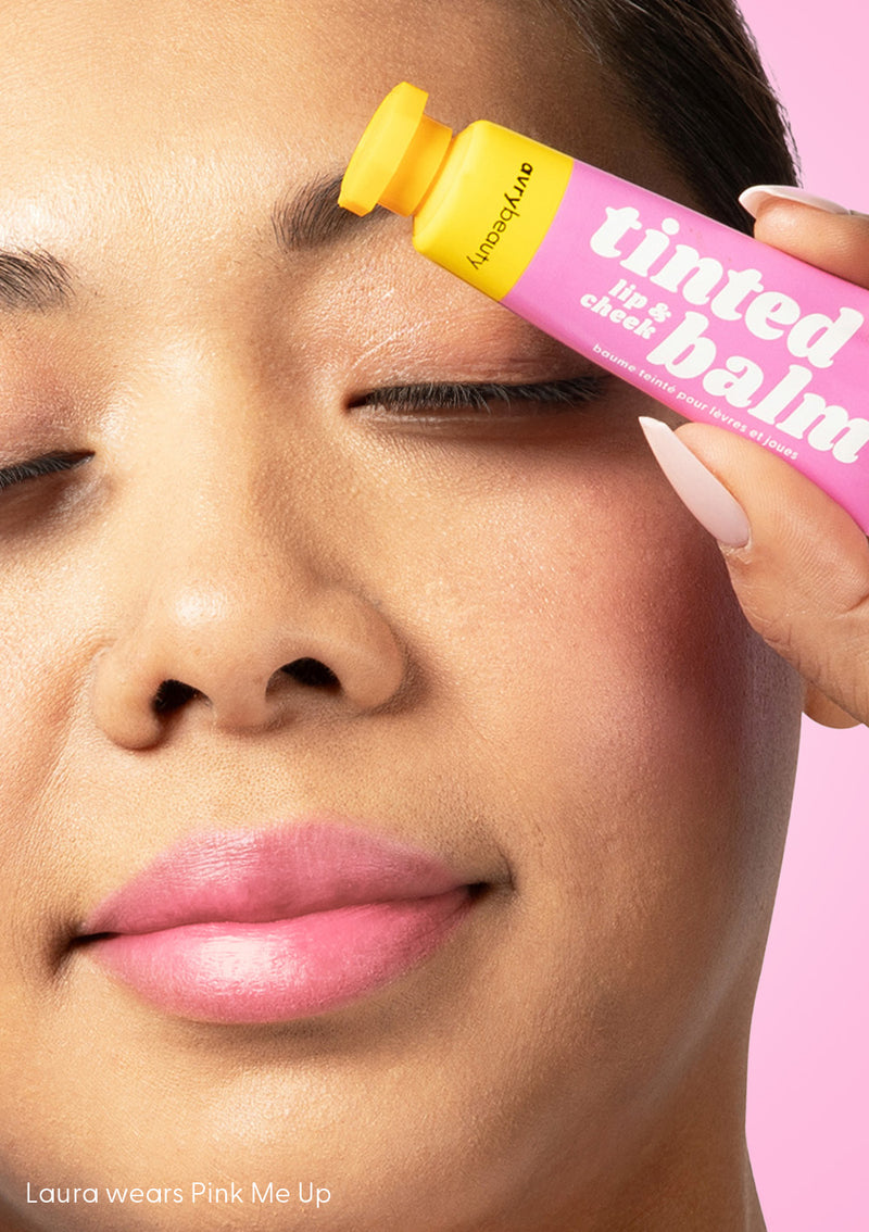 Laura wears AvryBeauty’s Pink Me Up Lip & Cheek Tinted Balm, a vegan makeup essential, on her lips, cheeks, and eyelids, while standing before a pink backdrop with eyes closed.
