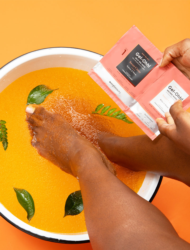 Female soaking feet in Sweet Citrus Gel-Ohh! jelly pedicure spa bath against an orange backdrop.
