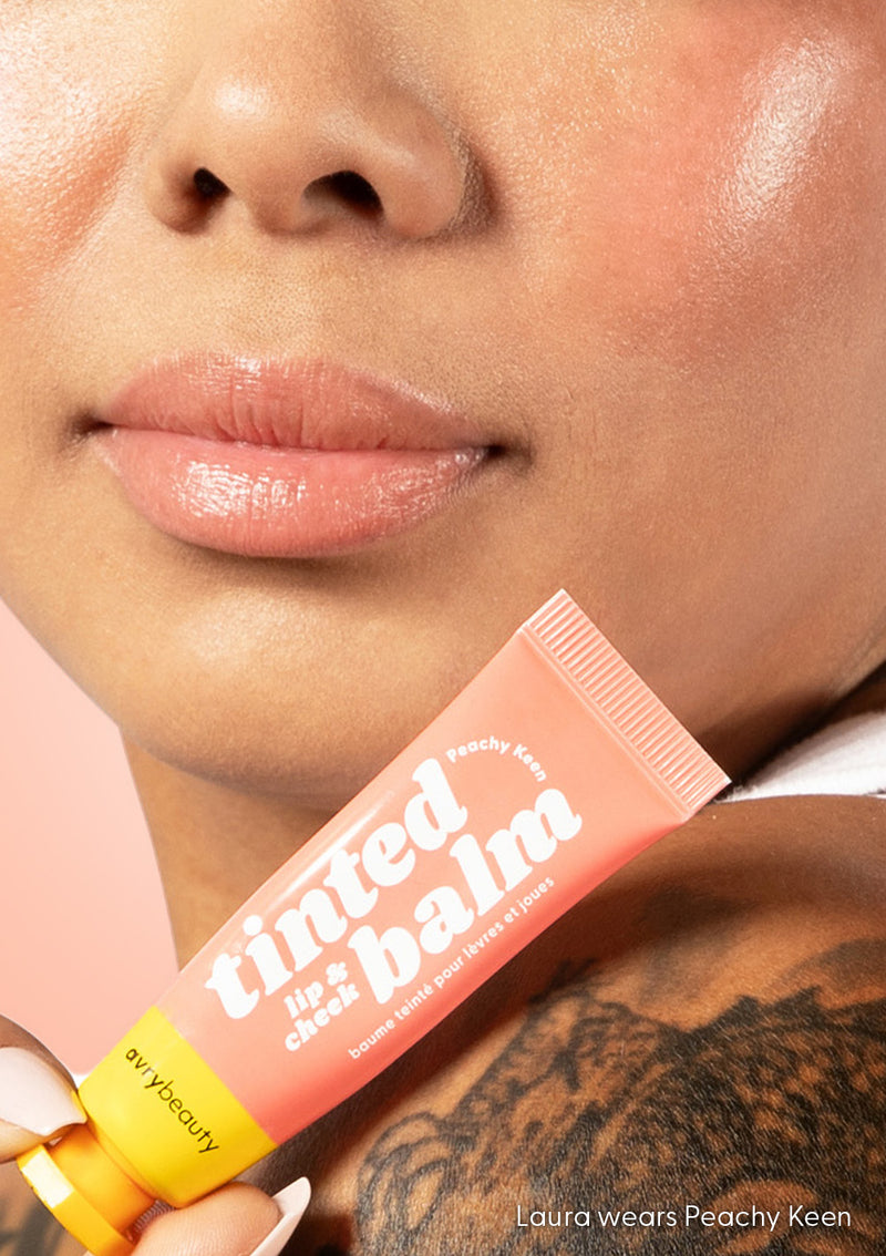 Laura standing in front of a peach-colored background, while holding up a tube of AvryBeauty’s Peachy Keen Lip & Cheek Tinted Balm, with the vegan makeup product applied to her lips and cheeks.
