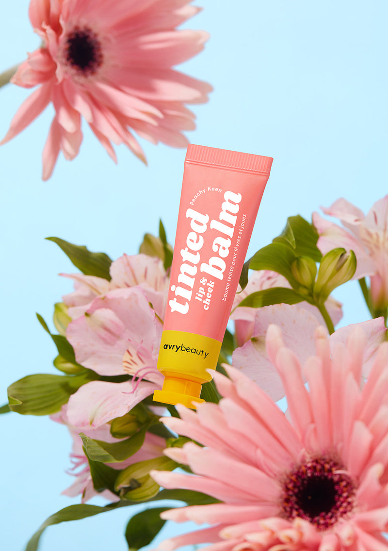 AvryBeauty’s Peachy Keen Lip & Cheek Tinted Balm, a 2-in-1 vegan skincare and makeup essential, placed on top of flowers with a blue background behind it.