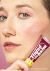 Bailey holds up a tube of Plum Delight Lip & Cheek Tinted Balm, with the 2-in-1 vegan makeup and skincare essential applied to her lips, cheeks, and eyelids, while standing in front of a purple background.