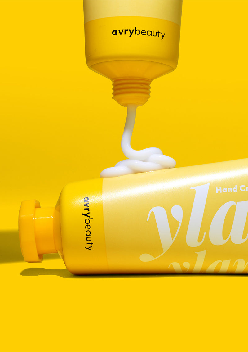 Ylang Ylang Shea Butter Lotion being squeezed out onto a second moisturizing hand cream bottle, set against a yellow background.